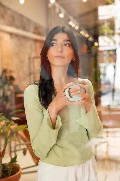 Femme pensive tenant une tasse, regard tourné vers le haut dans un espace chaleureux et végétalisé, illustrant la recherche de bien-être